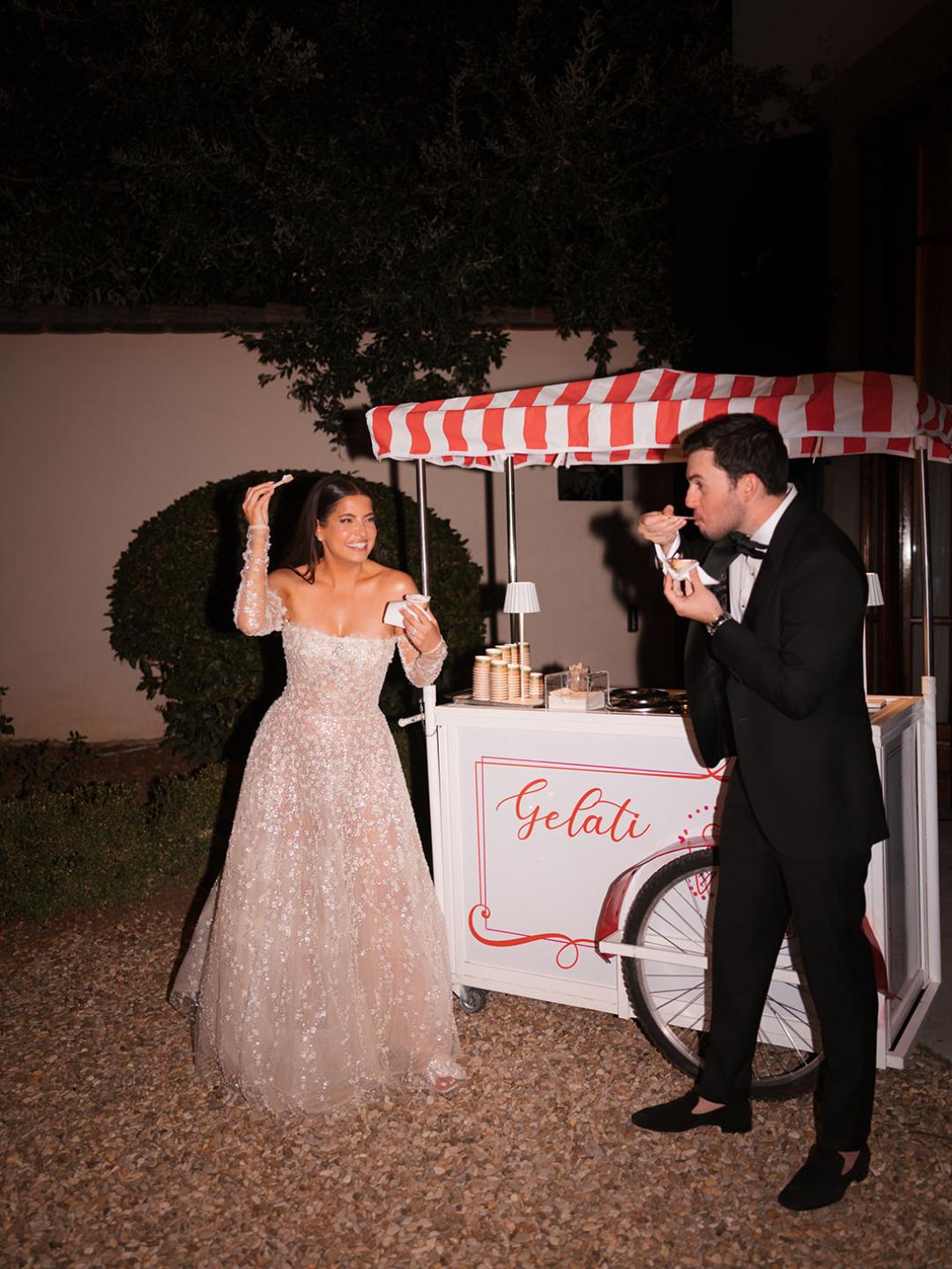 A woman in a wedding dress and a man in a tuxedo eat gelato in front of a small gelato cart at night.