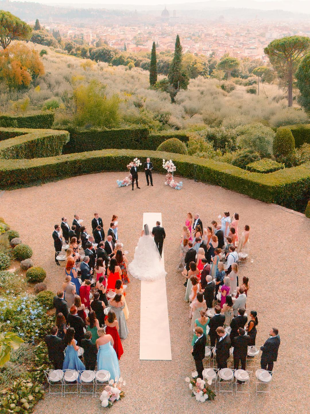 Outdoor wedding ceremony with guests seated on either side of a white aisle, bride and groom at the altar, and landscaped greenery in the background.