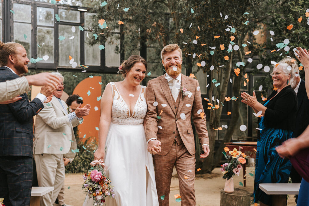 A bride and groom walk hand in hand down the aisle while guests throw confetti and applaud at an indoor wedding ceremony.