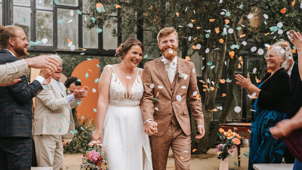 A bride and groom walk hand in hand down the aisle while guests throw confetti and applaud at an indoor wedding ceremony.