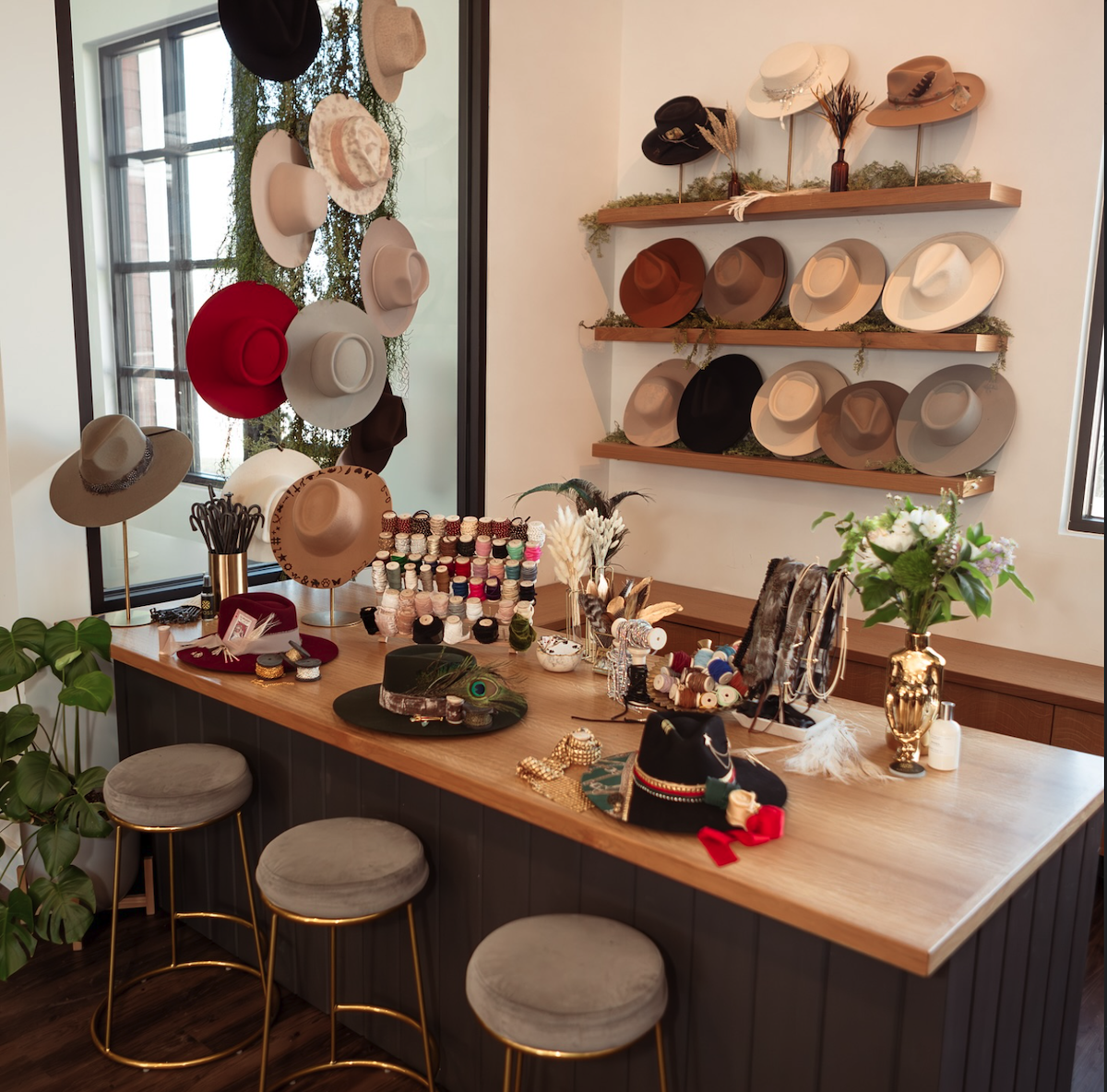 A boutique display features hats on walls and shelves, jewelry, hats, and accessories arranged on a wooden counter with three stools beneath.