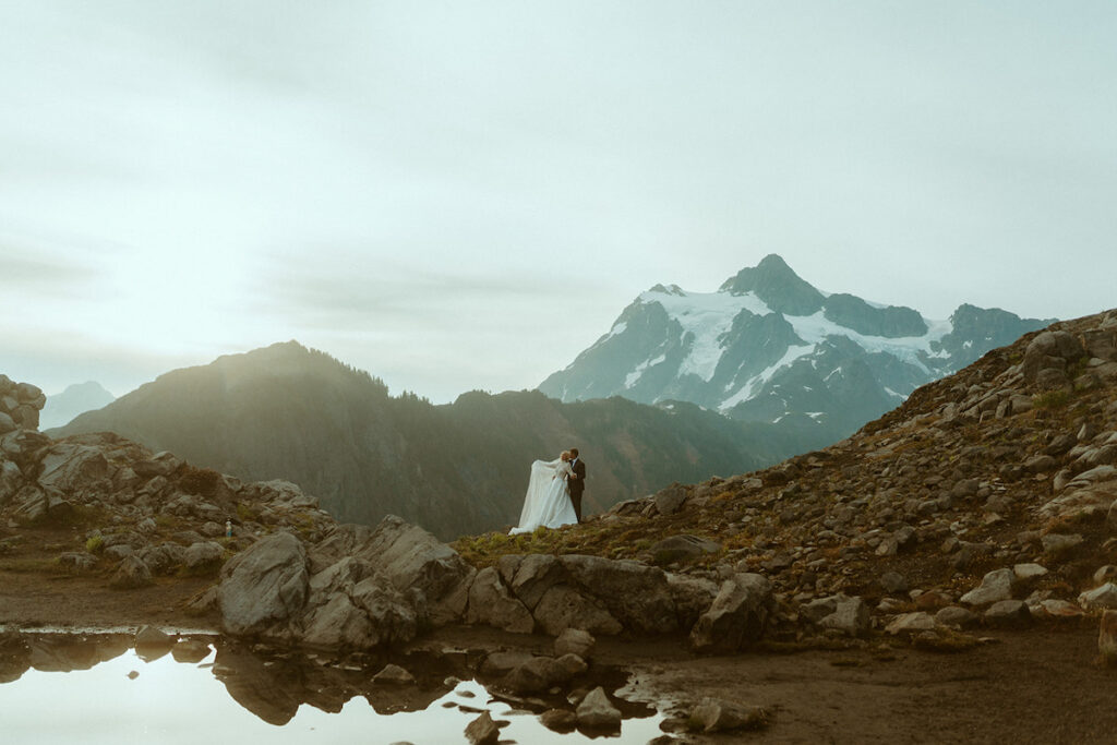 A couple in wedding attire stands on a rocky landscape with a snow-capped mountain and cloudy sky in the background.