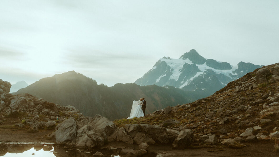 A couple in wedding attire stands on a rocky landscape with a snow-capped mountain and cloudy sky in the background.