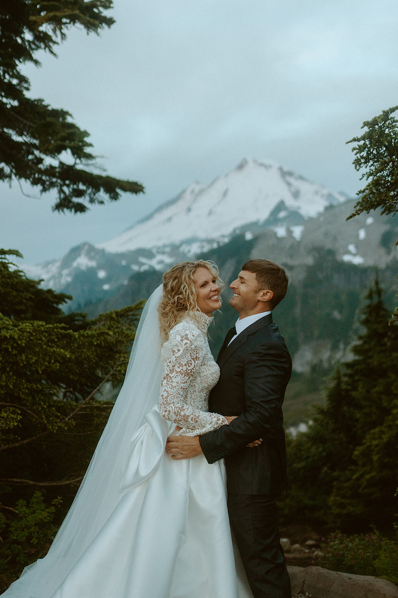 A bride and groom stand together outdoors in front of a snow-capped mountain, surrounded by trees.