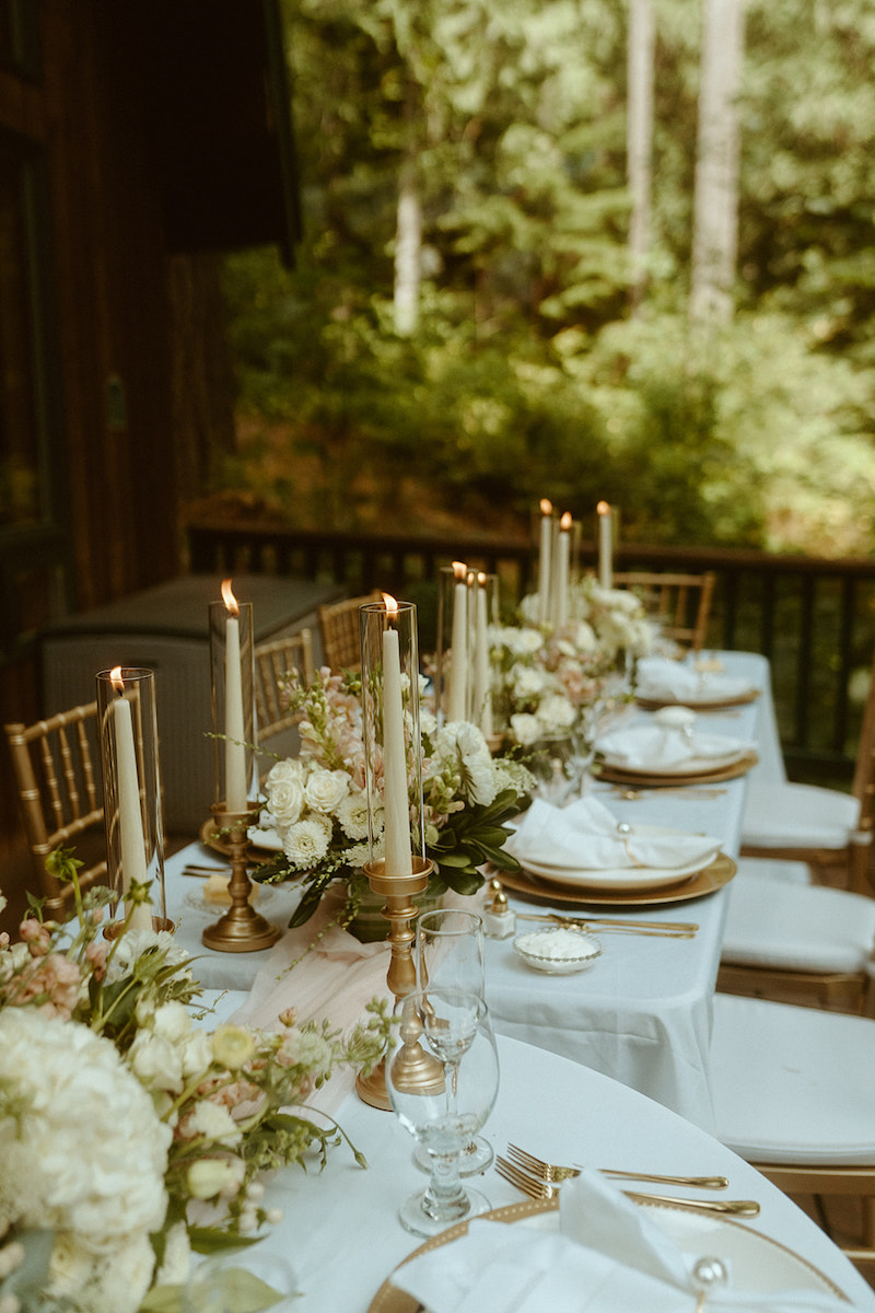 Elegant outdoor table setting with white linens, gold chairs, floral centerpieces, and tall candles, arranged for a formal event surrounded by greenery.