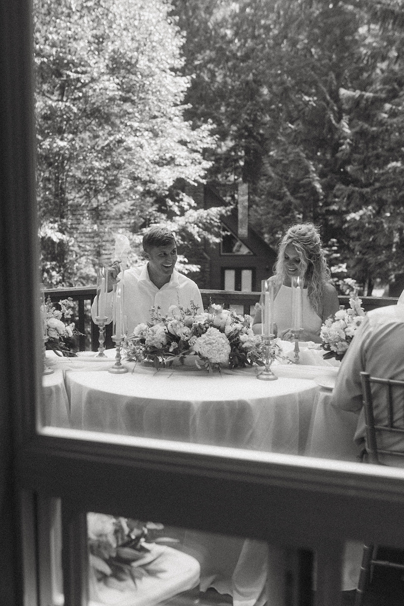 A black-and-white photo shows two people sitting at a round table outdoors, decorated with flowers and candles. Trees and a house are visible in the background through a window frame.