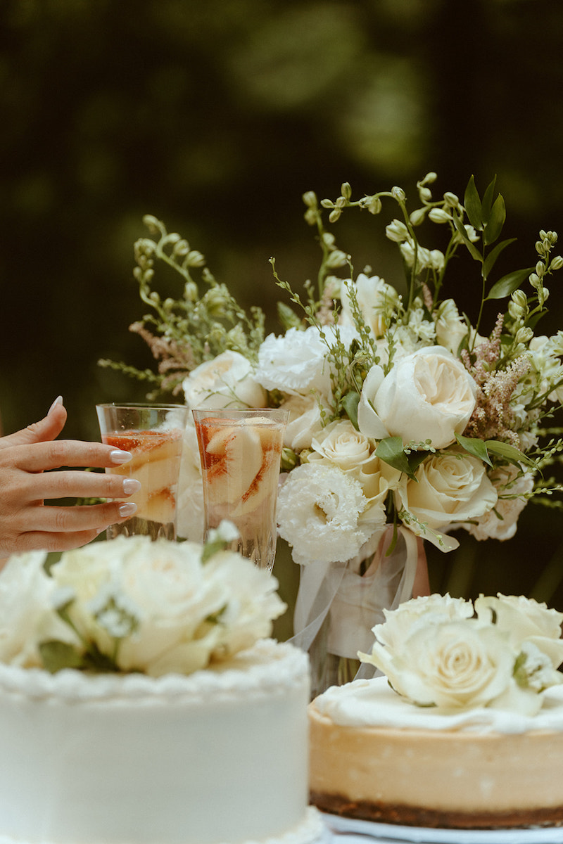 A hand holds a glass near a table with white rose flower arrangements and two cakes decorated with white roses.