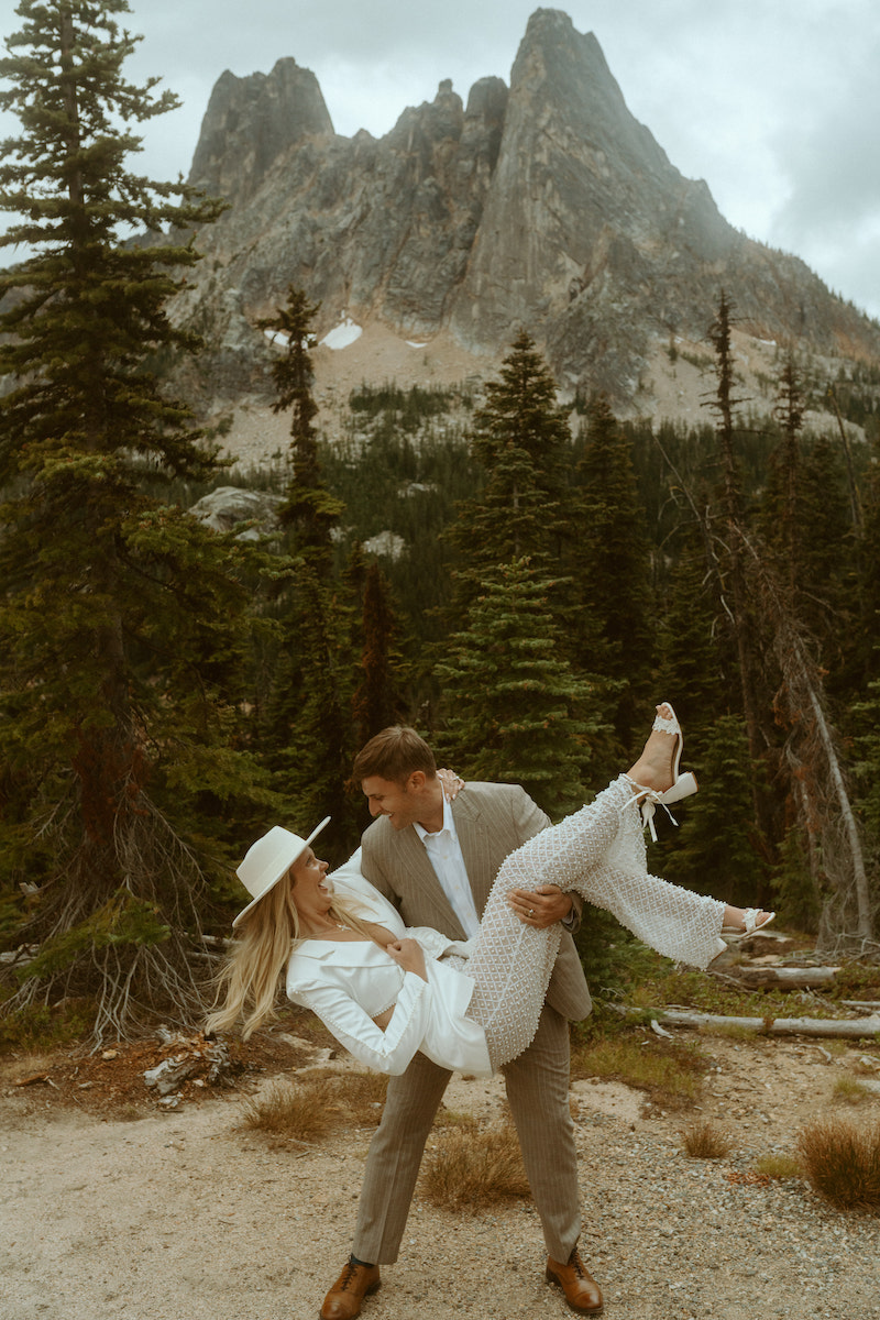A man in a suit holds a woman dressed in white and wearing a hat, with a mountainous landscape and pine trees in the background.