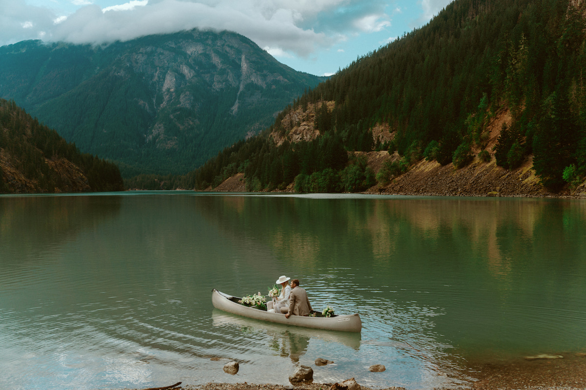 Two people in a canoe float on a calm lake surrounded by mountains and trees under a cloudy sky.