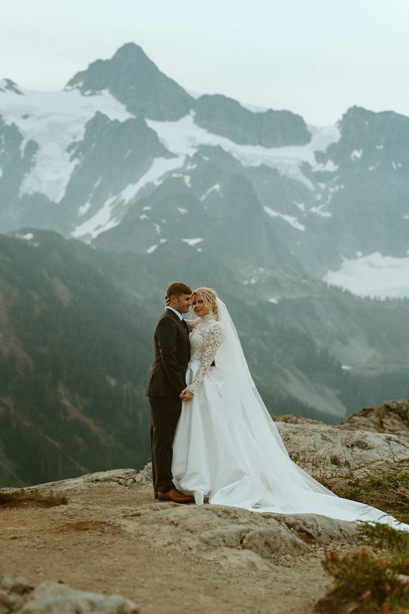 A bride and groom stand together on a rocky ledge with snow-capped mountains and cloudy sky in the background.