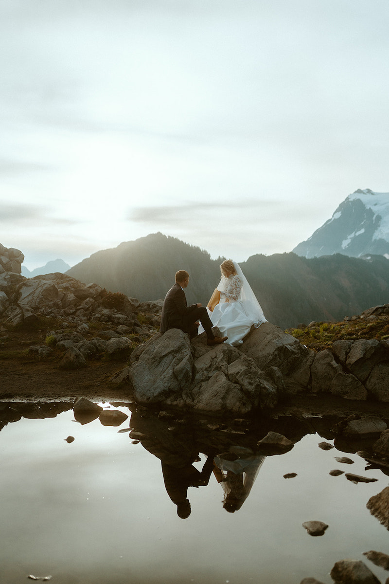 A bride and groom pose on rocks beside a small pool of water with mountain peaks in the background at sunset.