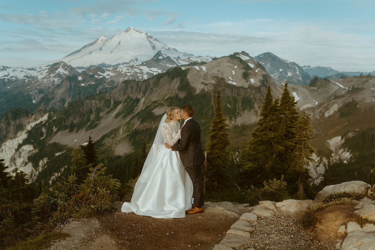 A bride and groom kiss on a mountain overlook with snow-capped peaks and evergreen trees in the background.