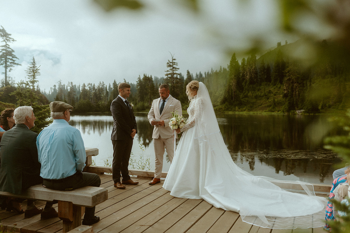 A bride and groom stand at a lakeside dock with an officiant, exchanging vows in front of seated guests surrounded by trees and water.