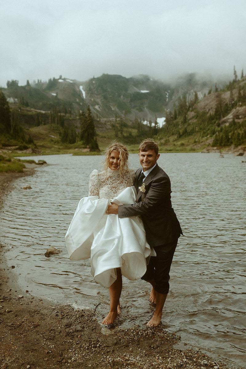 A groom in a suit carries a bride in a white dress while standing barefoot in a lake, with mountains and trees in the background.