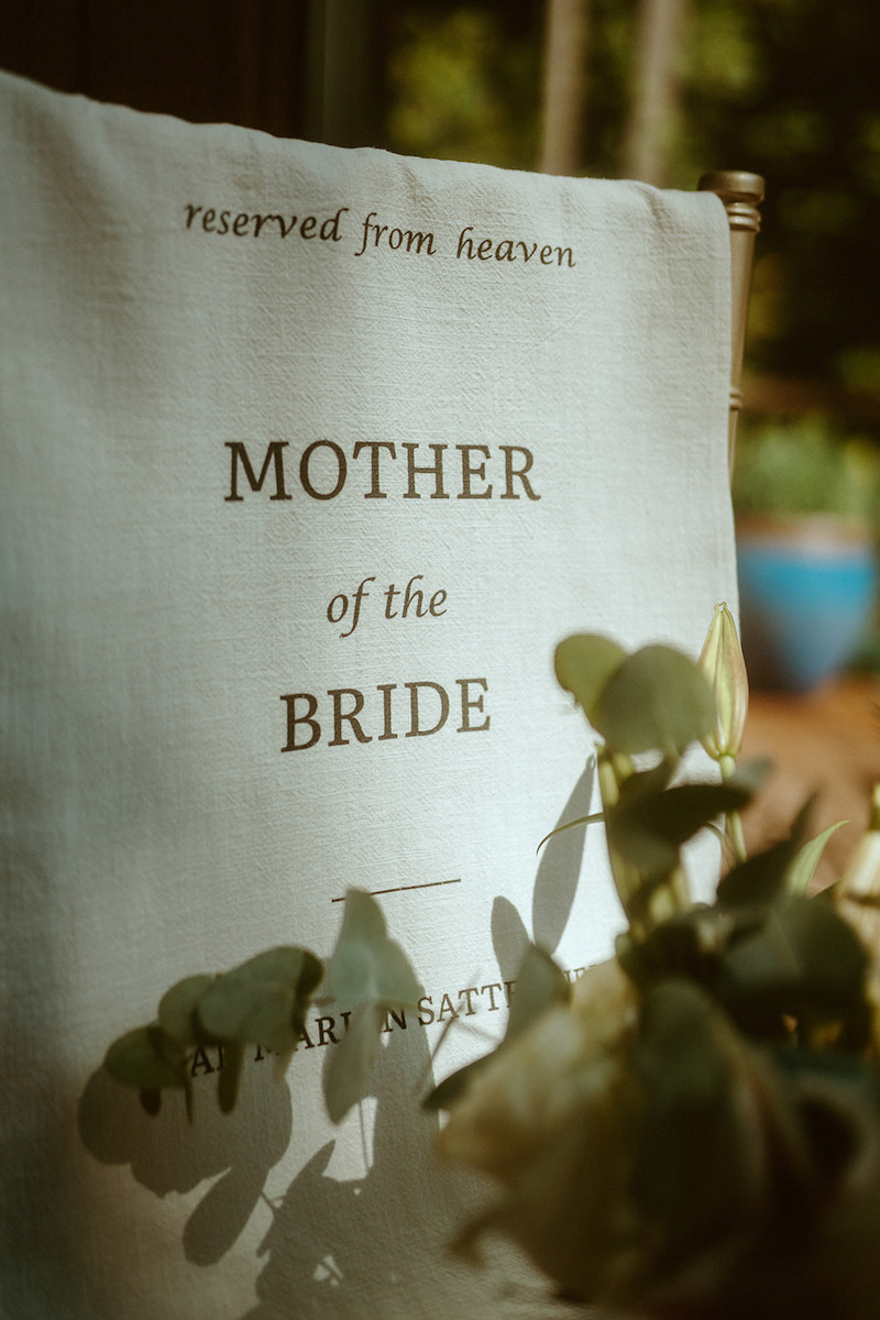 A white chair cover with the words "reserved from heaven, MOTHER of the BRIDE" printed on it, partially obscured by greenery in the foreground.