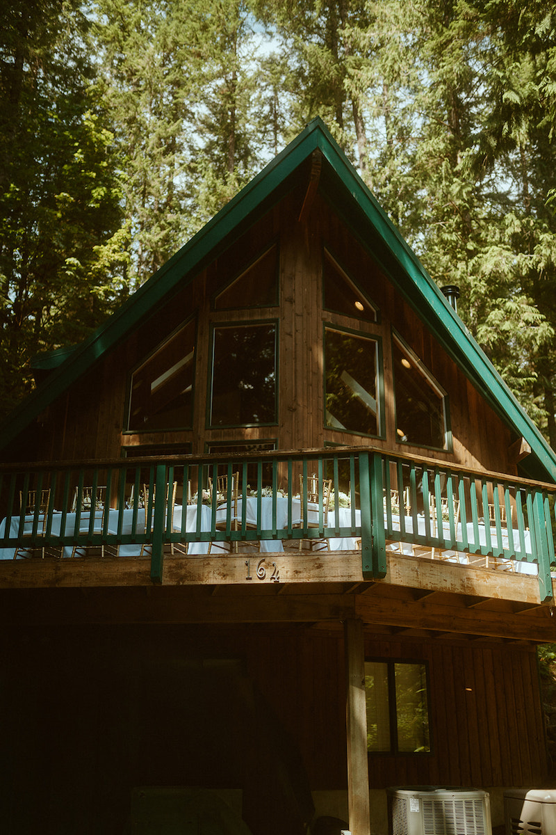 A wooden cabin with large windows and a green railing is elevated among tall trees in a forest setting.