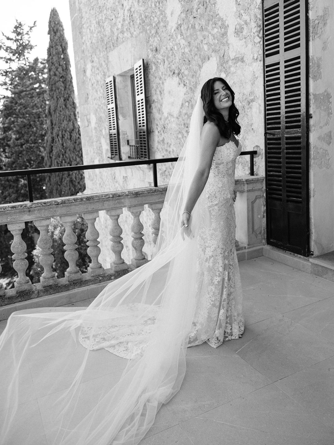 A bride in a strapless wedding dress with a long veil stands on a balcony, smiling and posing for the camera.