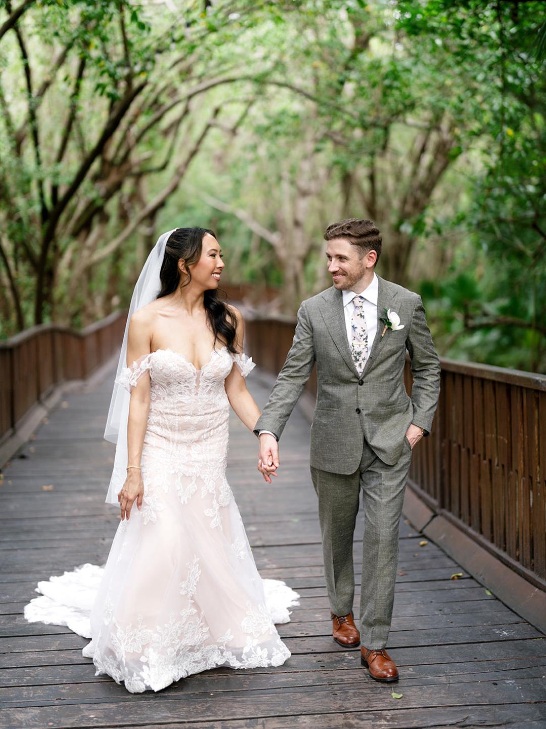 A bride in a white dress and a groom in a gray suit walk hand in hand on a wooden bridge surrounded by greenery.