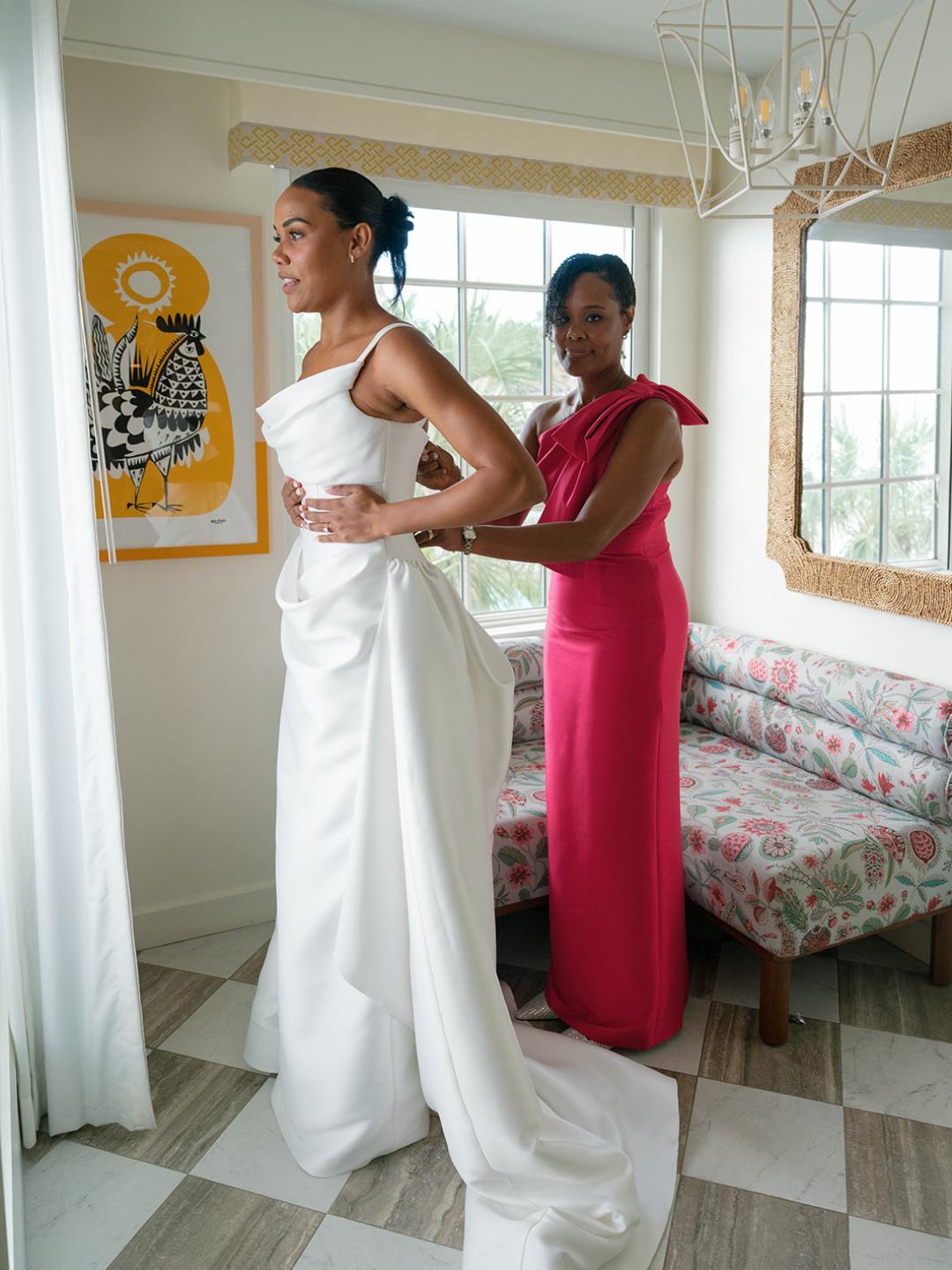 A woman in a white wedding dress stands near a window while another woman in a pink dress helps adjust the gown.