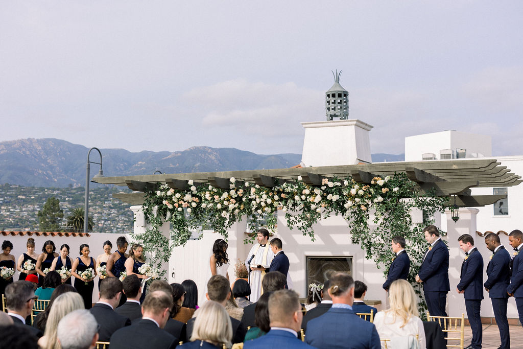 A wedding ceremony is taking place outdoors under a floral-decorated pergola, with guests seated and mountains visible in the background.