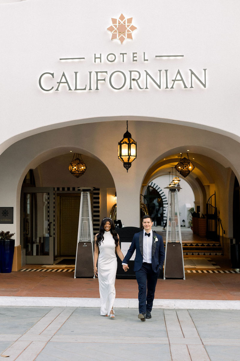 A couple holding hands walks under the archway entrance of the Hotel Californian, dressed in formal attire.