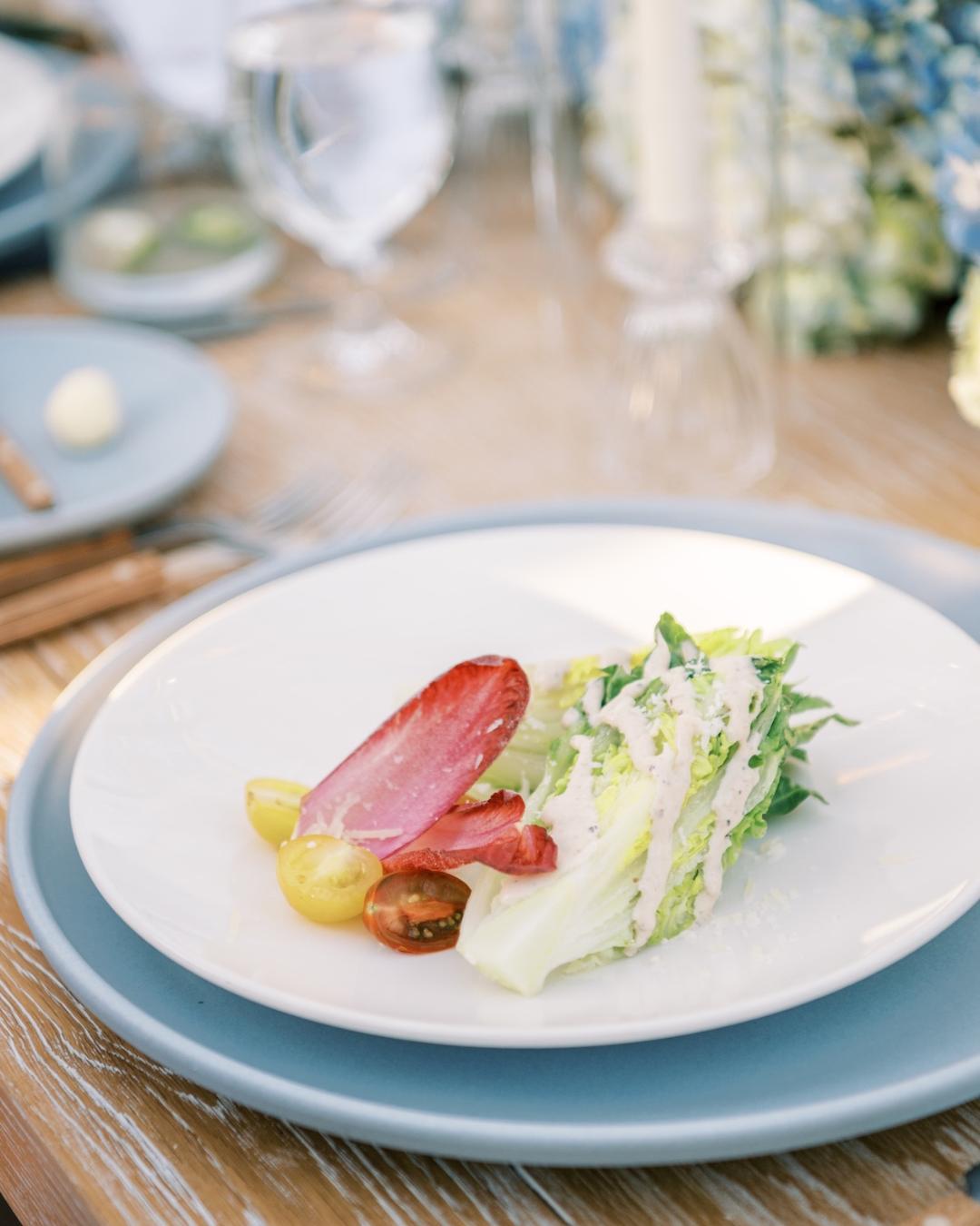 A plated salad with lettuce, grape tomatoes, and endive on a white dish set on a blue charger, with glassware and tableware in the background.