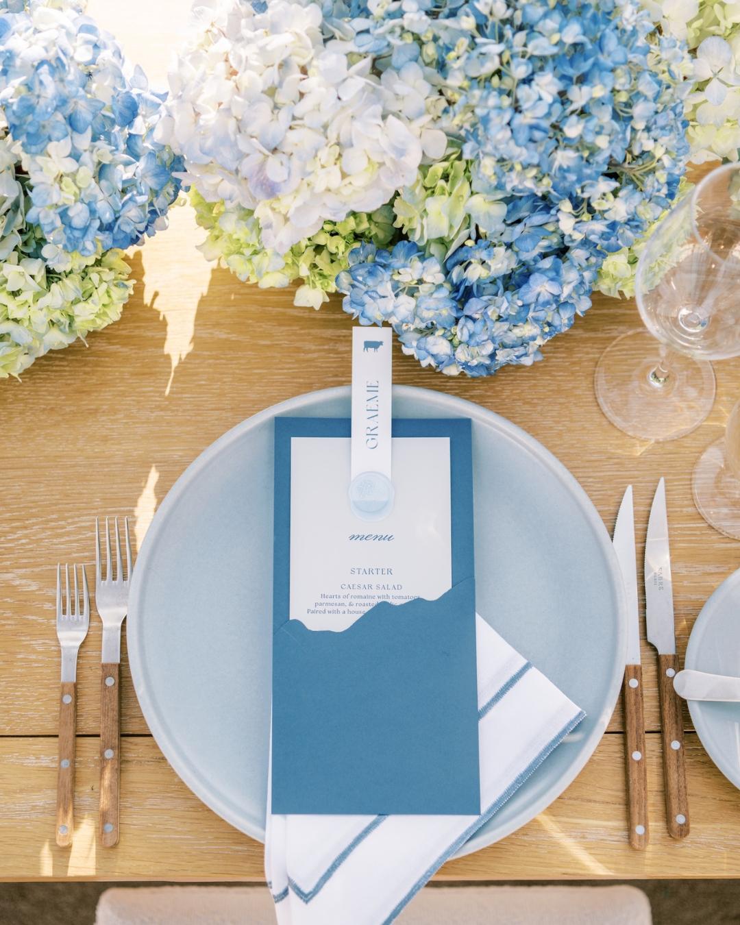 A blue and white place setting with a menu, napkin, and cutlery on a wooden table, surrounded by large blue and white hydrangea flower arrangements.
