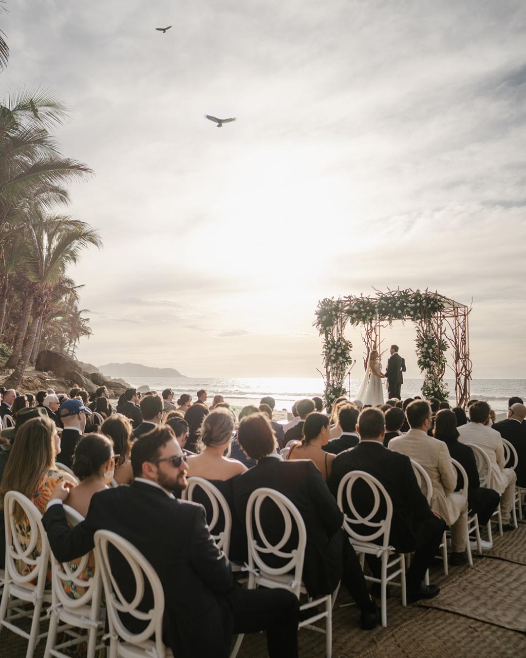 A group of guests sit facing a wedding altar on a beach at sunset, with two people standing under a floral arch and birds flying in the sky above.