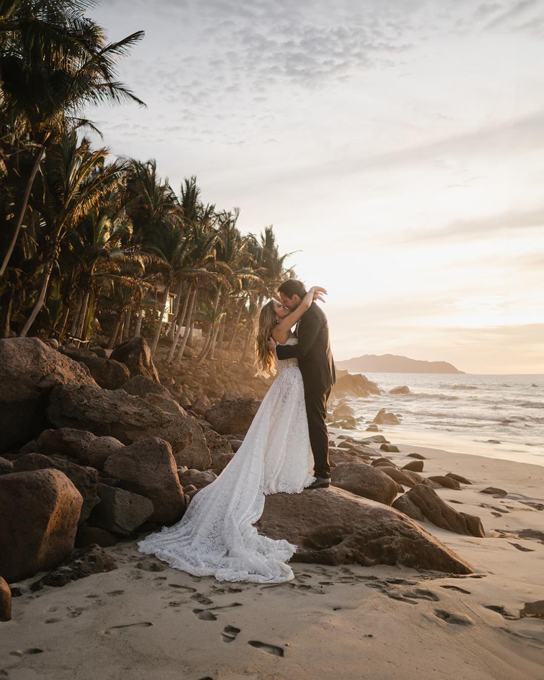 A bride and groom embrace on a rocky beach at sunset, with palm trees and ocean waves in the background.