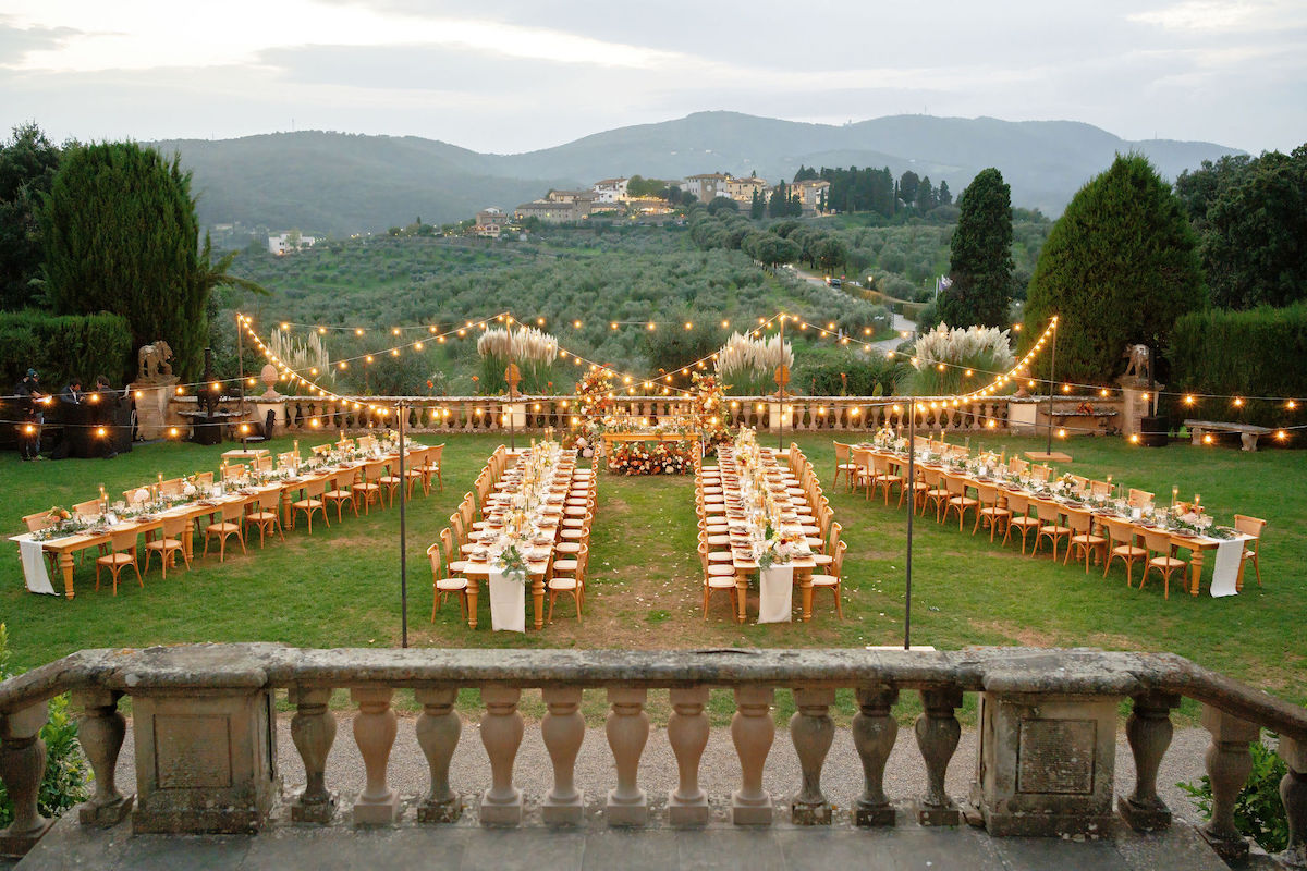 An outdoor formal dining setup with long tables, wooden chairs, and string lights, arranged on a lawn with hills and buildings visible in the background.