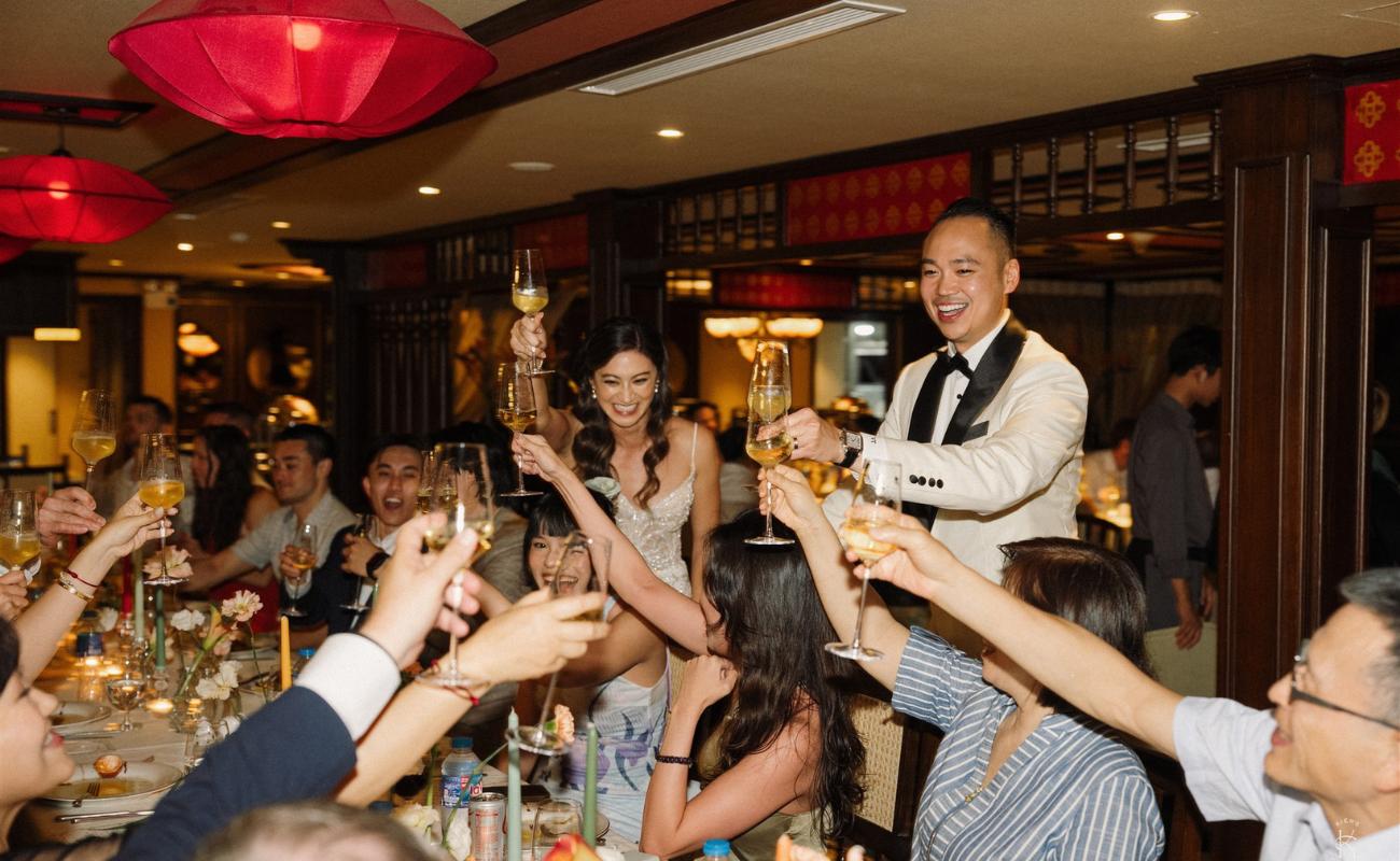 A group of people seated at a long table raise their glasses in a toast at a formal indoor celebration, with a couple standing and leading the toast.