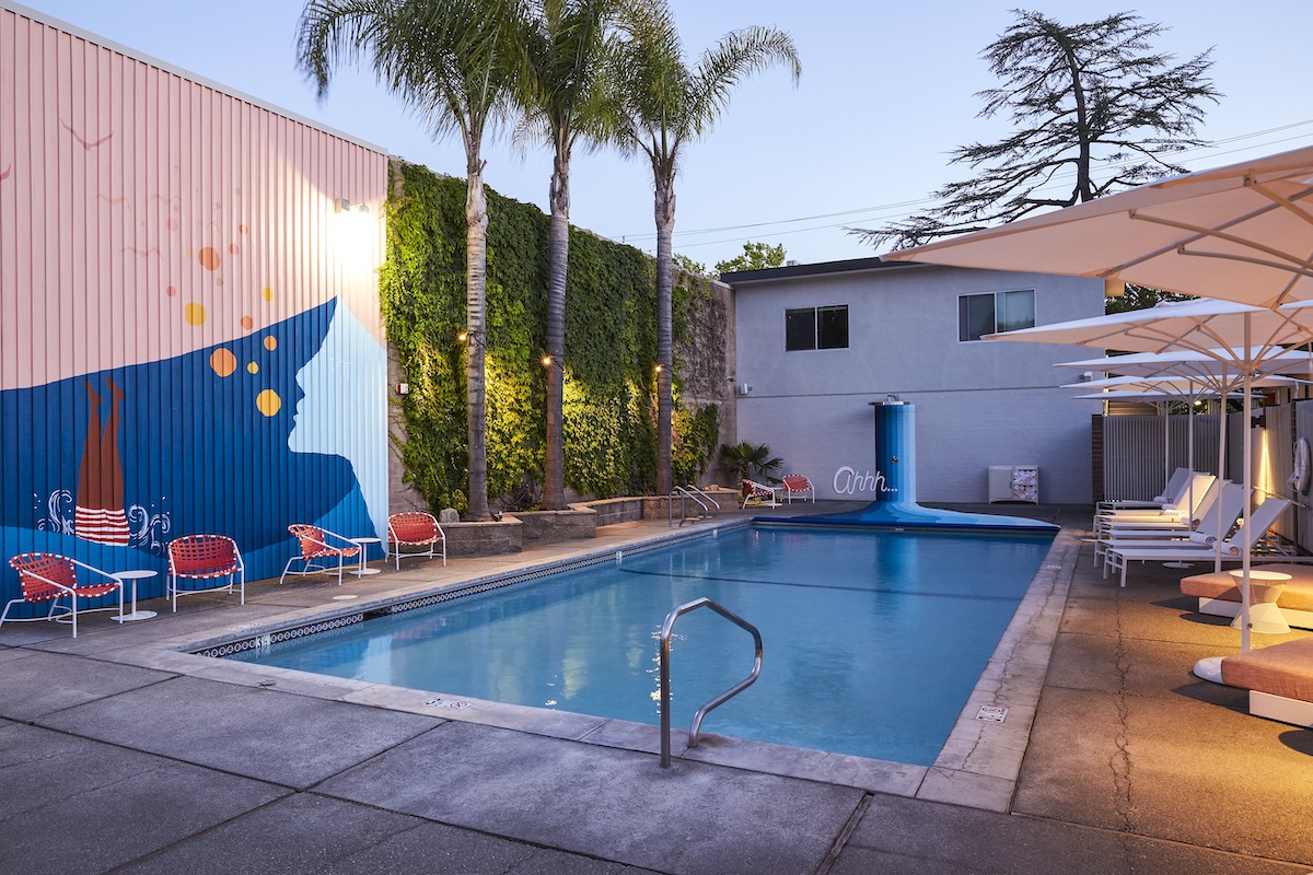 Outdoor swimming pool area with lounge chairs, umbrellas, palm trees, and a mural on the wall at dusk.