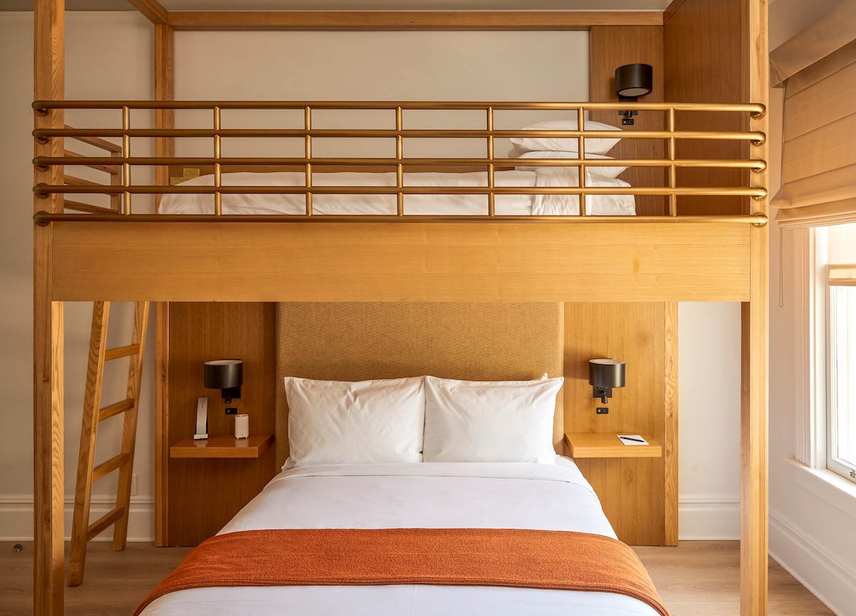 A room with a double bed below and a bunk bed above, both with white bedding and orange throws, wooden frame, bedside tables, lamps, and a window to the right.