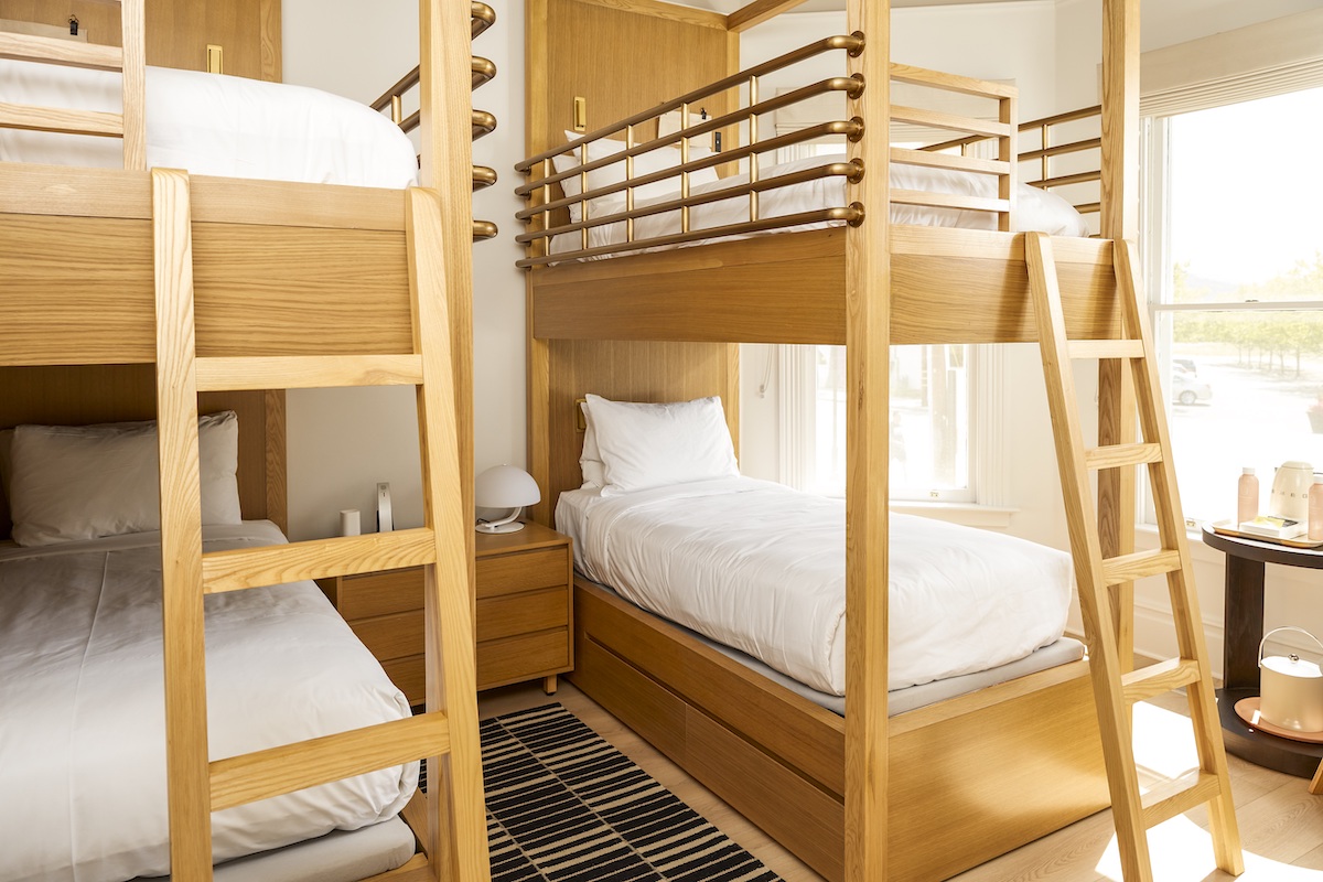 A bright bedroom featuring two wooden bunk beds, white bedding, a small nightstand with a lamp, and a window letting in natural light.