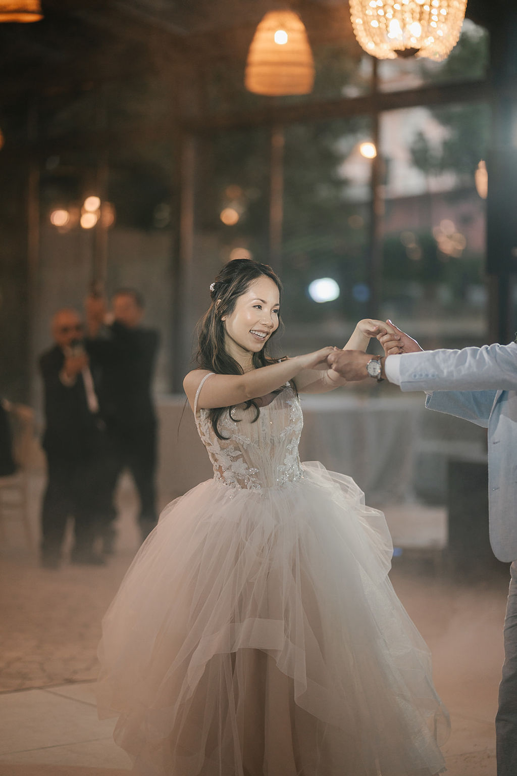 A bride in a white wedding dress dances with a partner indoors, surrounded by soft lighting and a misty atmosphere.