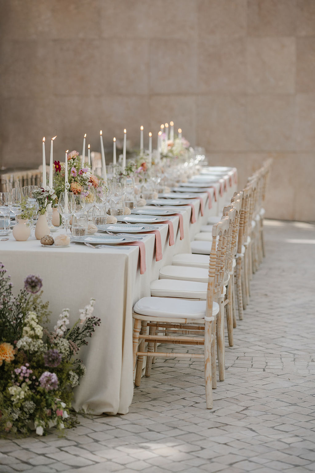 A long, elegant dining table is set outdoors with white tablecloth, candles, flowers, and neatly arranged chairs, ready for a formal event or celebration.