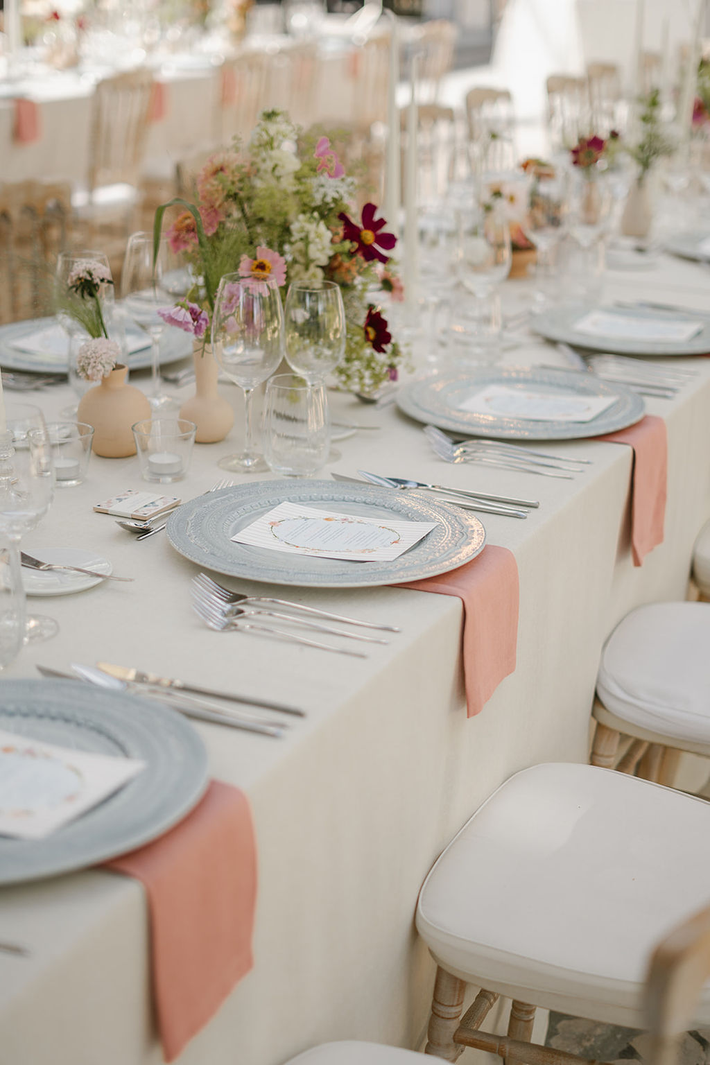 Elegant table setting with white tablecloth, light blue plates, pink napkins, floral centerpieces, and glassware arranged for a formal event or celebration.