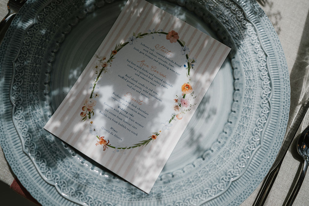 A printed menu with floral designs rests on a decorative blue plate, with shadow patterns cast across it.