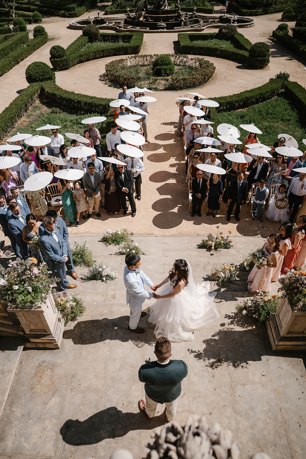 A couple stands at the altar holding hands during an outdoor wedding ceremony, surrounded by guests holding white parasols in a formal garden setting.