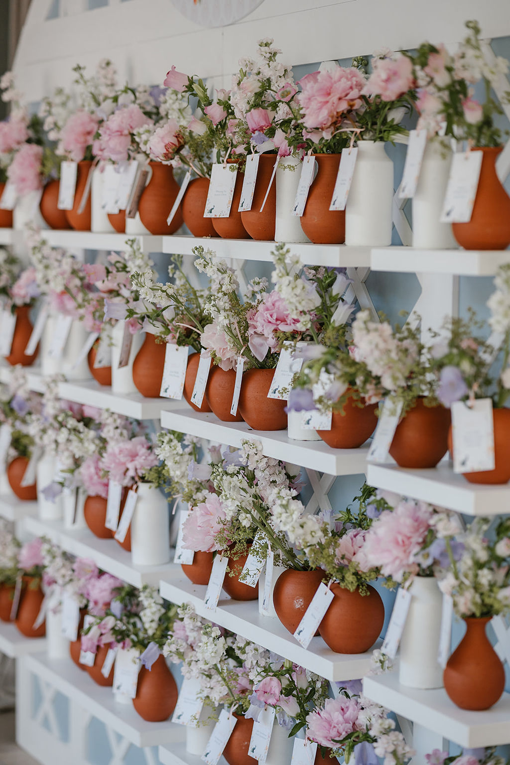 Rows of brown and white pitchers filled with pink and white flowers are arranged on white shelves, each pitcher labeled with a tag.