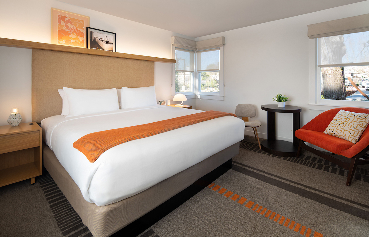 Modern hotel room with a neatly made king bed, orange throw, bedside tables, armchair, small round table with chairs, and large windows letting in natural light.