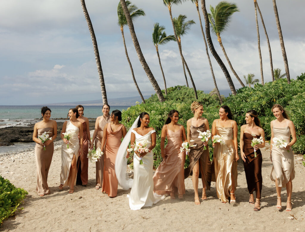 A bride in a white dress walks on a beach with eight bridesmaids in assorted neutral dresses, all holding bouquets, with palm trees and ocean in the background.