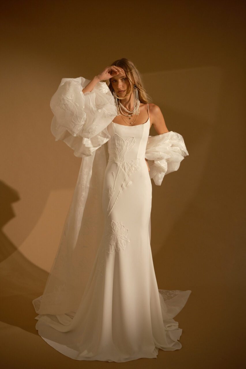 A woman wearing a long, white, sleeveless gown with sheer ruffled sleeves stands against a plain, softly lit brown background.