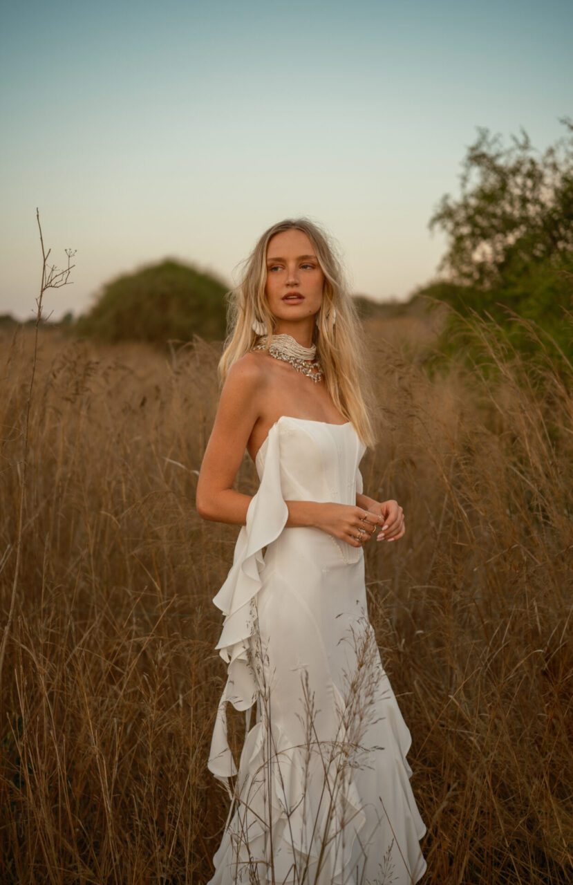 A woman in a strapless white dress stands in a dry grassy field at sunset, looking slightly away from the camera with greenery in the background.