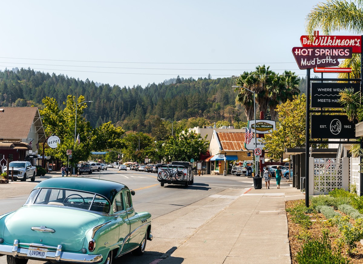 A vintage turquoise car is parked on a small town street with shops, palm trees, and a sign for Dr. Wilkinson’s Hot Springs in the background.