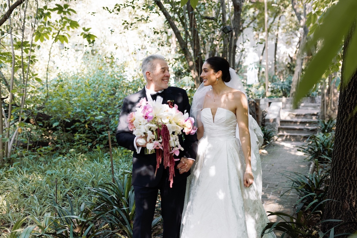 A bride in a white dress walks outside with an older man in a tuxedo holding a bouquet; they are smiling at each other on a garden path.