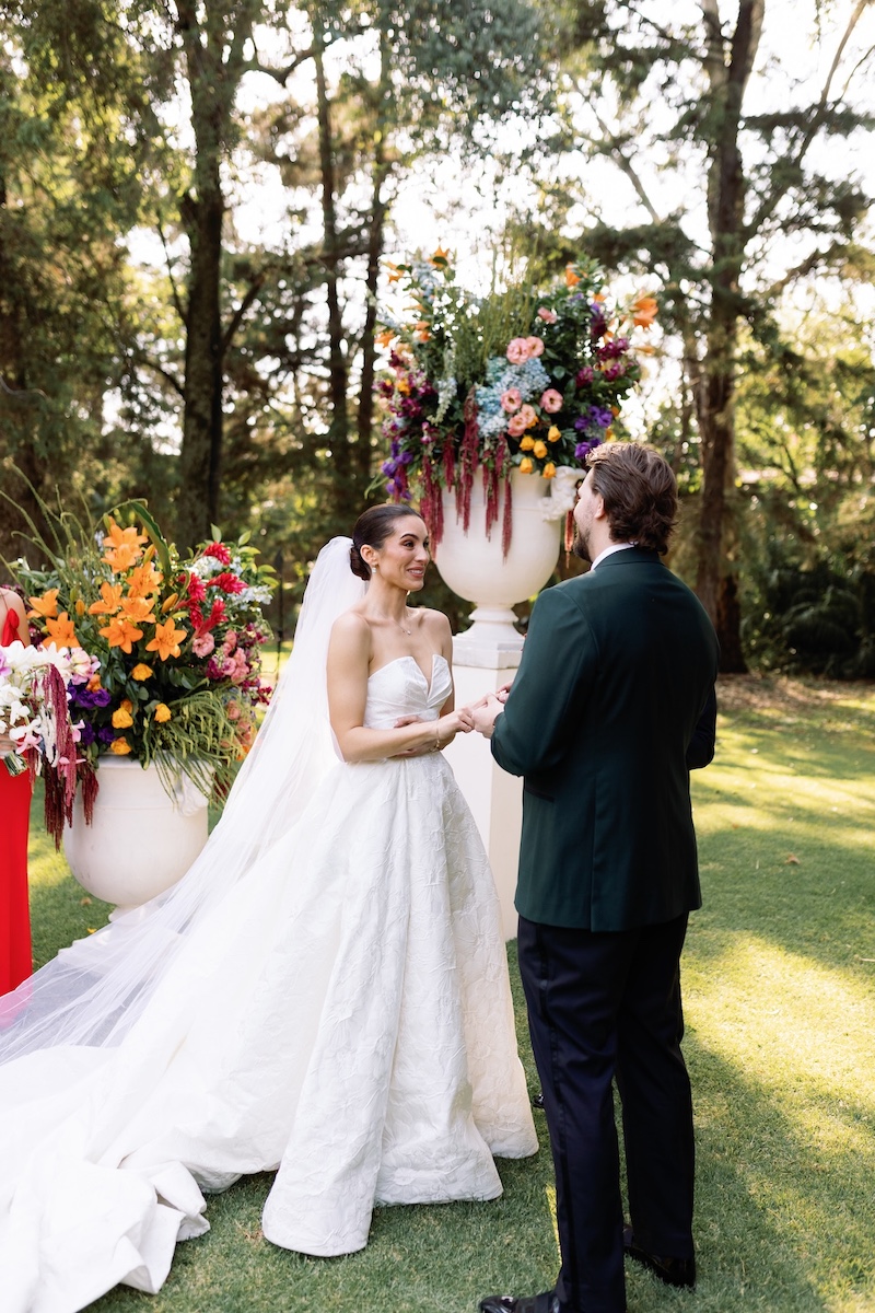 A bride and groom stand outdoors exchanging vows, surrounded by large floral arrangements and trees in the background.
