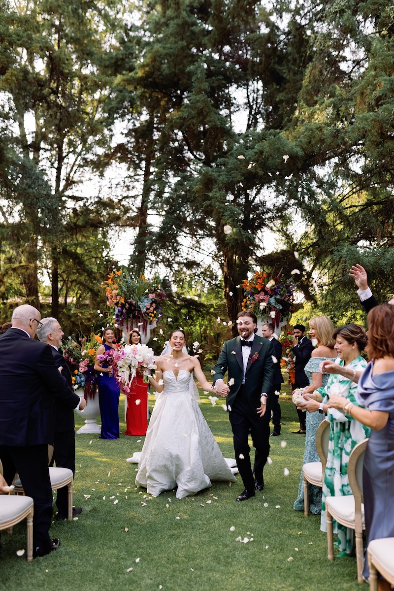 A bride and groom walk down an outdoor aisle, smiling and holding hands, as guests celebrate and throw flower petals.