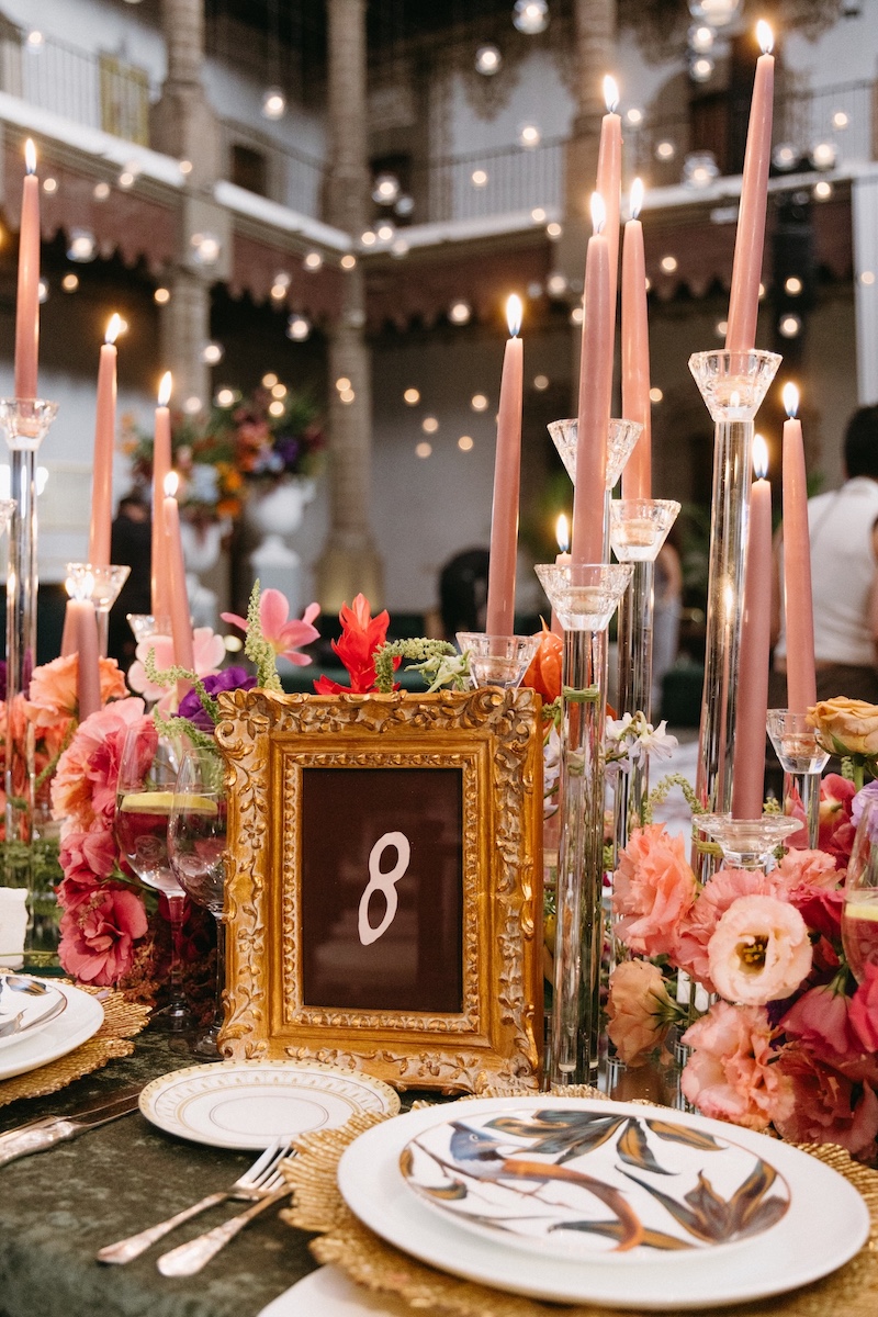 Ornate table setting with pink candles, colorful flowers, plates, and a gold-framed table number eight, under string lights in an indoor venue.