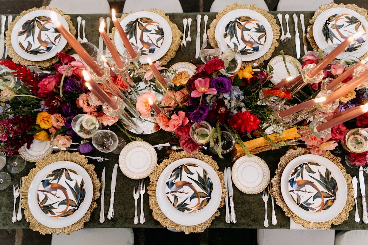 A formal dining table set with bird-themed plates, gold-rimmed chargers, assorted cutlery, tall candles, and colorful floral centerpieces.